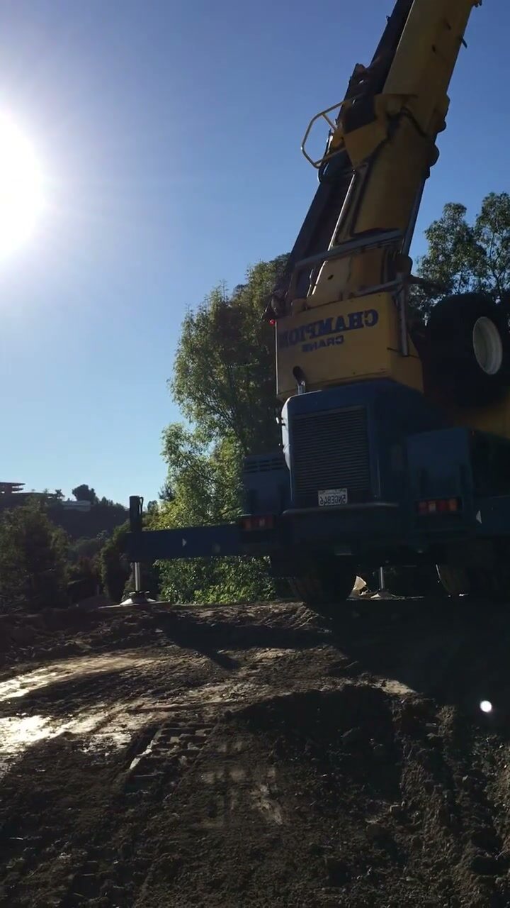 A crane-mounted drill rig silhouetted against the sun on a hillside excavation site.