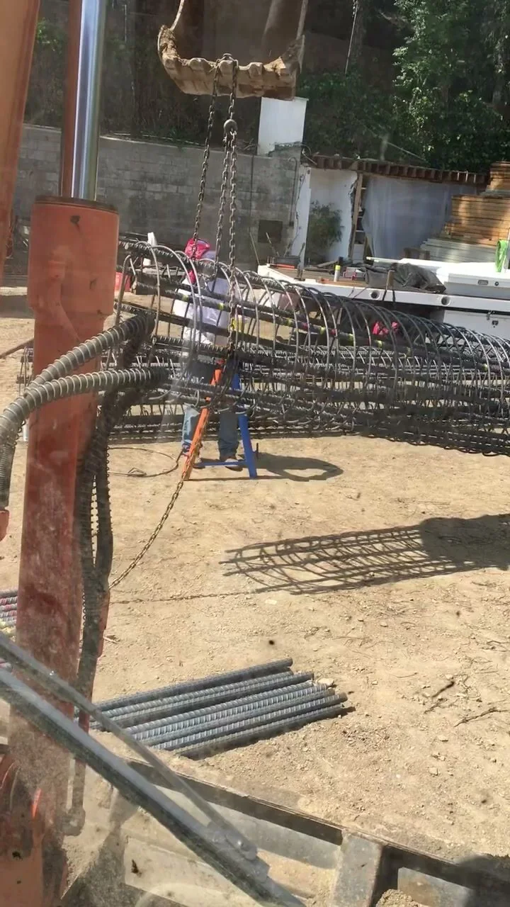 An excavator bucket lowering a large steel rebar cage into place at a retaining wall footing.