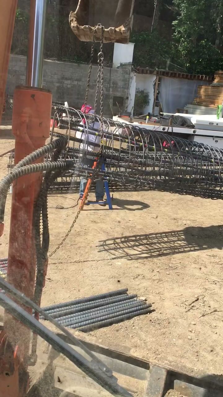 An excavator bucket lowering a large steel rebar cage into place at a retaining wall footing.