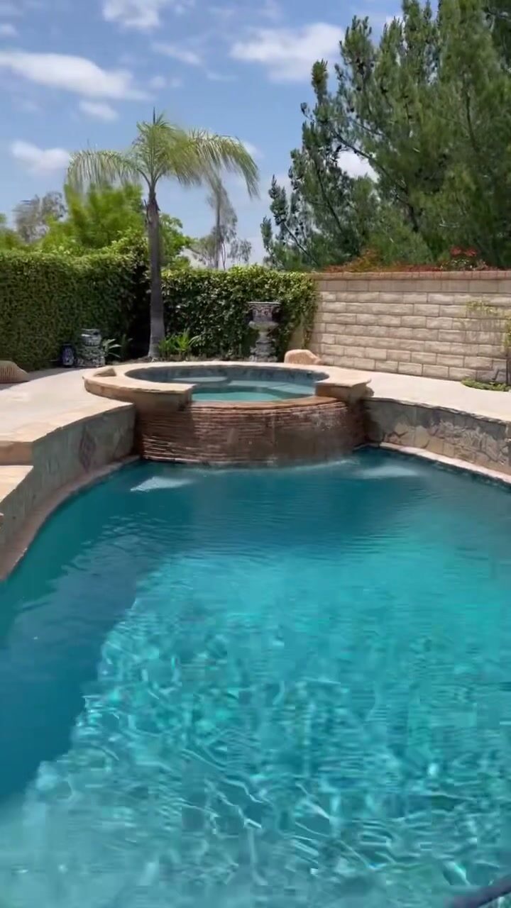 A finished backyard pool and raised spa with a stacked-stone waterfall, framed by mature palm trees and a manicured hedge — a Sierra Development hardscape build.